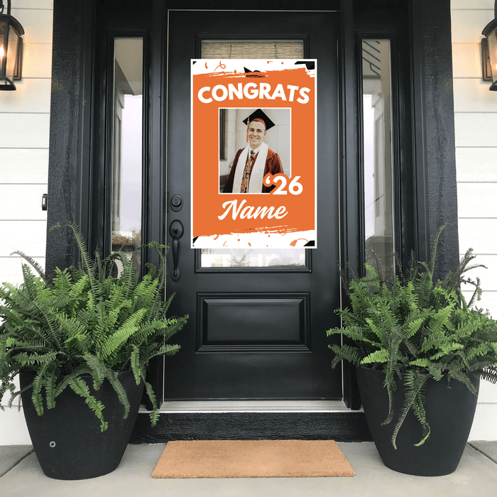 Graduation-themed poster on a black door with potted plants on either side.