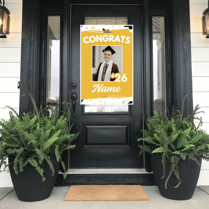 Graduation-themed personalized poster on a black door with potted plants and a doormat.