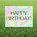 Holographic happy birthday yard sign featuring an iridescent pastel rainbow background, thin black “Happy Birthday!” lettering, and a white rectangular border displayed in grass.