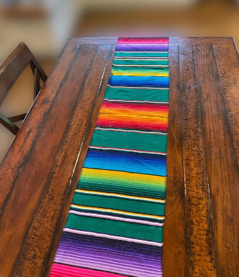 Mexican serape table runner featuring bright multicolor woven stripes in pink, purple, blue, yellow, red, orange, and green displayed down the center of a wooden table.