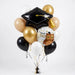 Graduation balloon bouquet featuring a black graduation cap-shaped foil balloon with a gold tassel, a round “Congrats Grad” owl foil balloon, and black, white, gold, and tan latex balloons with curling ribbons.